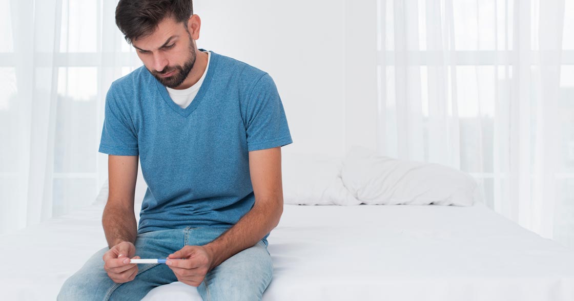 man sitting on a bed holding a pregnancy looking despondent